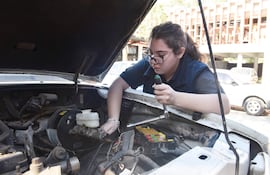 Mujer de cabello castaño con gafas trabaja en un motor de auto utilizando una herramienta, en un ambiente exterior tranquilo.