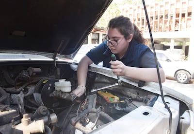 Mujer de cabello castaño con gafas trabaja en un motor de auto utilizando una herramienta, en un ambiente exterior tranquilo.
