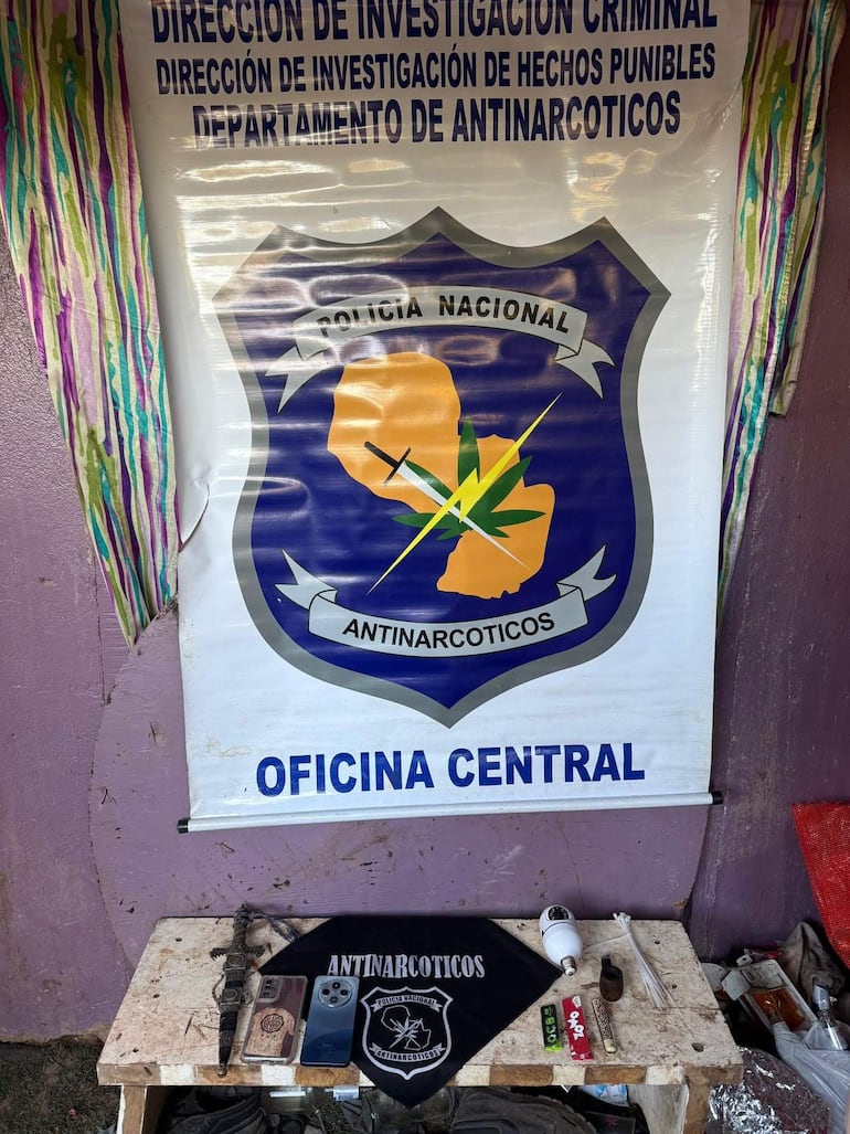 Espacio de trabajo con cartel de Antinarcóticos, móvil, documentos y objetos vinculados al narcotráfico sobre una mesa.