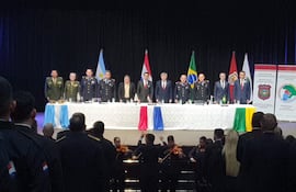 Tres hombres en uniforme, con expresiones tristes, observan desde una mesa con banderas de Argentina, Paraguay y Brasil al fondo.