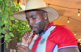 Yonathan Destine, con camiseta de Paraguay y sombrero de paja, sostiene un vaso mientras disfruta en un ambiente acogedor.