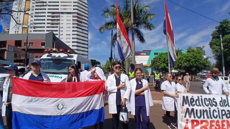 Grupo de médicos con batas blancas sostiene una gran bandera paraguaya y un cartel en una calle urbana con vegetación.