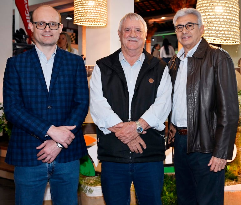 Tres hombres sonrientes: uno en blazer a cuadros, otro en chaleco negro y el tercero en chaqueta de cuero marrón.