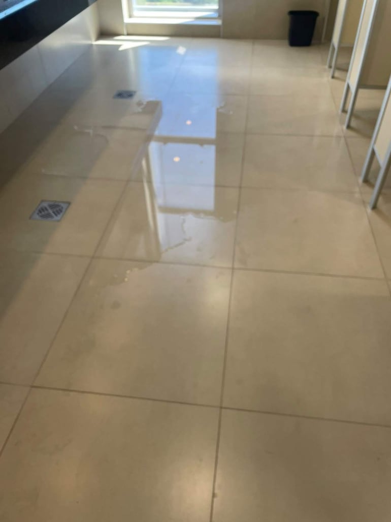 El agua filtra en los sanitarios y no alcanza para el uso de inodoros ni para el lavado de manos.
El ambiente se ve iluminado, debido a la luz natural que entra por una ventana ubicada al fondo a la derecha. No se identifican elementos que puedan relacionarse con un equipo de fútbol, como camisetas o escudos. 

No hay objetos relevantes adicionales en el contexto visual, como carteles o pantallas, que aporten información sobre el lugar o la situación. La imagen sugiere un posible problema de filtración o limpieza debido a las marcas de agua.