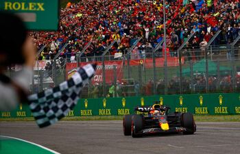 Max Verstappen en coche de Red Bull alcanzando la meta, con mano sosteniendo bandera a cuadros y audiencia animada en el fondo.