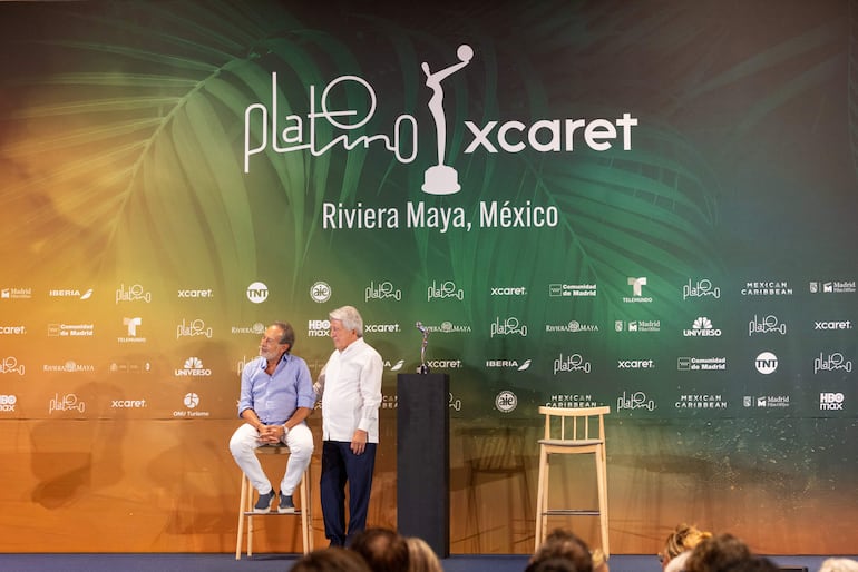 Guillermo Francella sentado en taburete con camisa azul, conversa con un hombre de pie con cabello blanco y camisa blanca en evento decorado.