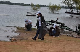 Cuatro personas en camisetas blancas con 'TEAM TAIWAN' recogen basura en la orilla de un río, con gorras blancas y un ambiente natural.