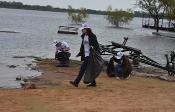 Cuatro personas en camisetas blancas con 'TEAM TAIWAN' recogen basura en la orilla de un río, con gorras blancas y un ambiente natural.