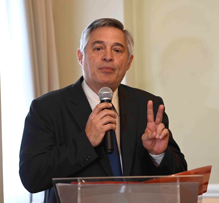Luis Ramírez, con cabello canoso y traje oscuro, sostiene un micrófono y gesticula con paz en un ambiente formal.