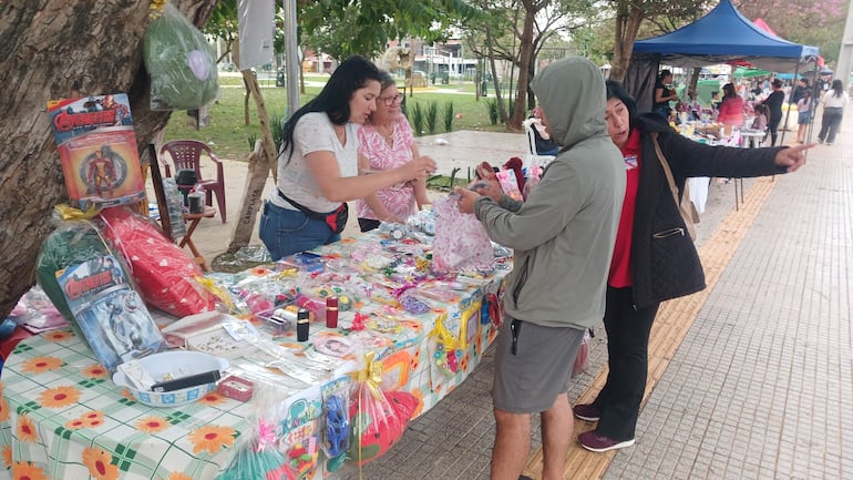 Mujeres en feria, una con cabello largo y oscuro conversando con un hombre en chaqueta gris, rodeadas de mesas con artículos diversos.