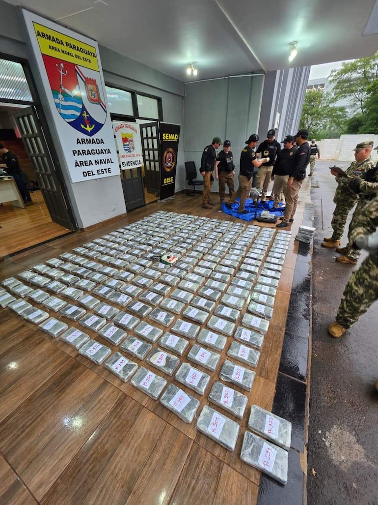 Seis hombres uniformados organizan paquetes etiquetados de drogas en el suelo, bajo un escudo de la Armada Paraguaya en un ambiente militar.
