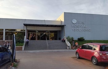 Dos mujeres suben escaleras del hospital, mientras un grupo conversa en la escalinata. Vehículos estacionados en el fondo.