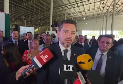 Pedro Alliana en traje oscuro habla a la cámara, acompañado de un hombre en traje claro y una multitud atenta detrás.