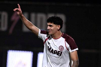 Jugador del Club Lanús con camiseta blanca y burdeos, cabello corto y rizado, sonríe levantando el dedo índice en un estadio oscuro.