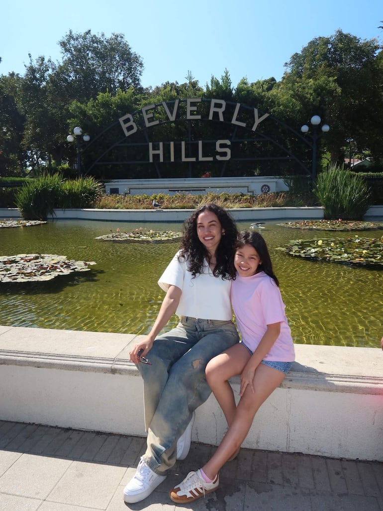 Mujer con cabello rizado y blusa blanca sonríe, junto a niña de camiseta rosa y pantalones cortos, ambos en un parque soleado.
