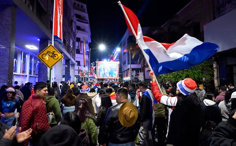 Grupo de personas con prendas rojas, blancas y azules, mirando atención hacia una pantalla grande en un ambiente festivo.