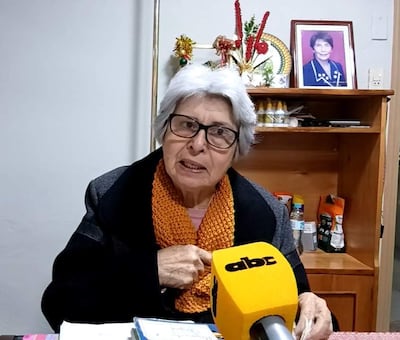 Corina Falcón, mujer mayor con gafas y bufanda naranja, habla expresivamente frente a un micrófono amarillo en un ambiente acogedor.