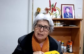 Corina Falcón, mujer mayor con gafas y bufanda naranja, habla expresivamente frente a un micrófono amarillo en un ambiente acogedor.