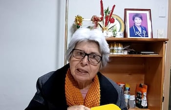 Corina Falcón, mujer mayor con gafas y bufanda naranja, habla expresivamente frente a un micrófono amarillo en un ambiente acogedor.