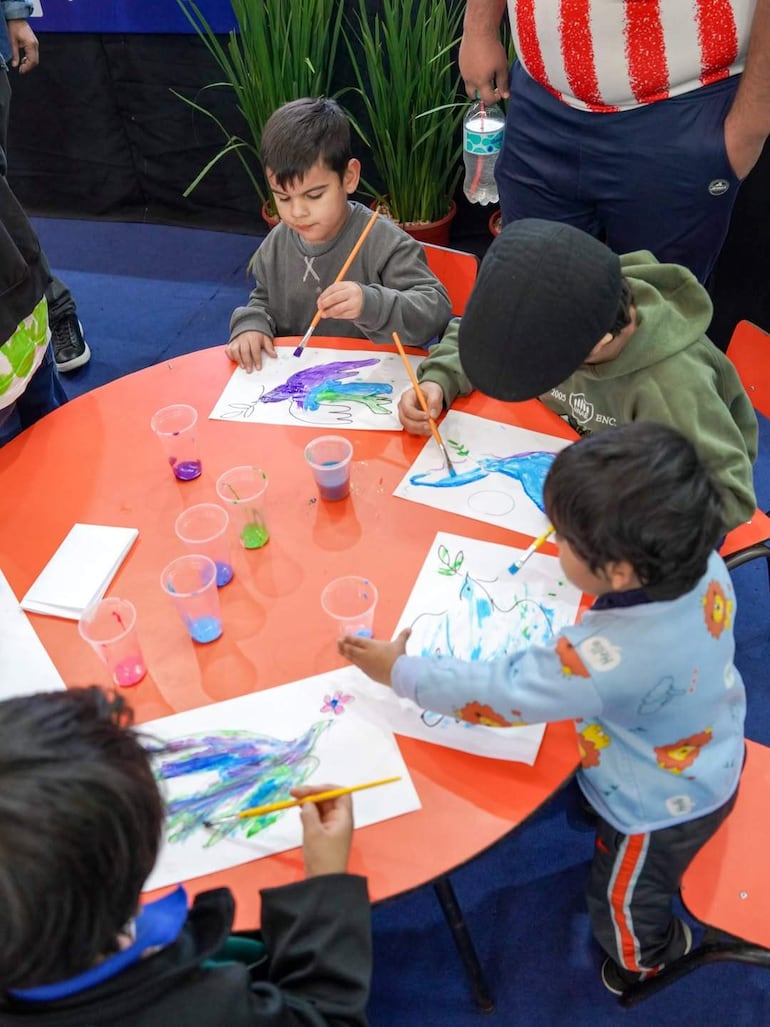 Cuatro niños concentran en sus dibujos con papeles y colores, en ambiente colorido y supervisados por un adulto.