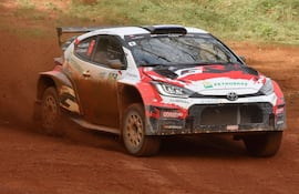 Automóvil de rally Toyota salpicando tierra en un camino, con espectadores observando en el fondo.
