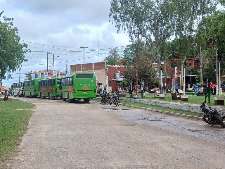Buses verdes estacionados en una calle, con un entorno urbano lleno de áreas verdes y personas en bancos, mientras llueve.