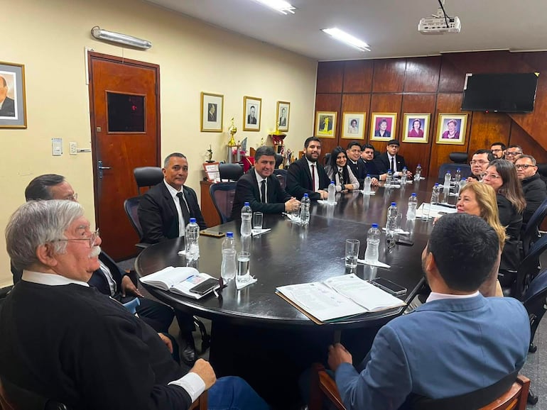 Grupo de 16 personas en reunión formal, algunos hombres y mujeres con trajes oscuros, ambiente de oficina, atención en la conversación.