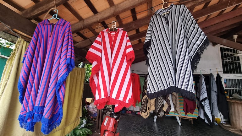 Tres ponchos con rayas, uno morado y azul, otro rojo y blanco, y otro negro y blanco, exhibidos en un ambiente iluminado.