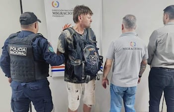 Un oficial de policía y tres hombres en el interior de una oficina. Uno, con camiseta negra y shorts, es asistido por funcionarios de migraciones.