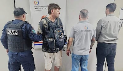 Un oficial de policía y tres hombres en el interior de una oficina. Uno, con camiseta negra y shorts, es asistido por funcionarios de migraciones.