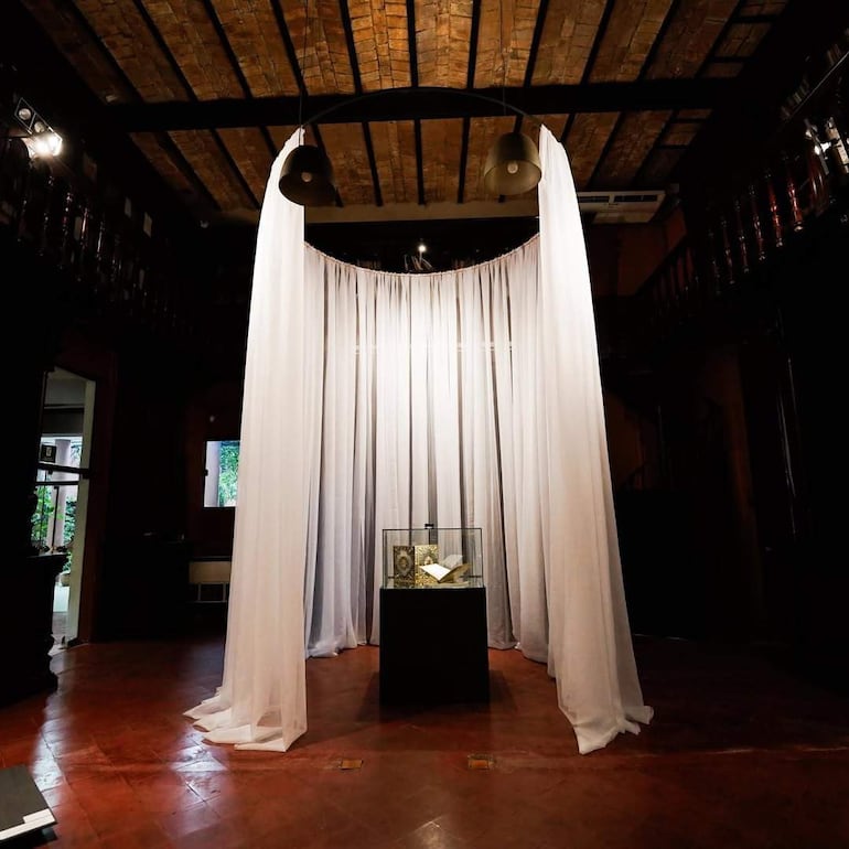 Vitrina con libro antiguo en pedestal negro, rodeada de telón blanco en un espacio tranquilo con suelo de cerámica.