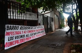 Persona con chaqueta oscura camina por acera mojada, junto a un banner que denuncia la situación del barrio Las Mercedes.