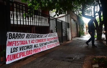 Persona con chaqueta oscura camina por acera mojada, junto a un banner que denuncia la situación del barrio Las Mercedes.