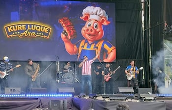 Cinco músicos actúan en el escenario, el vocalista en camiseta a rayas, bajo el humo festivo, con pantalla de fondo colorida.