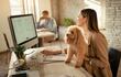Joven empresaria feliz trabajando en una computadora mientras está con su perro en la oficina.