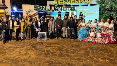 Grupo de 20 personas en trajes tradicionales coloridos sonríen con arco de bienvenida al fondo durante evento festivo.