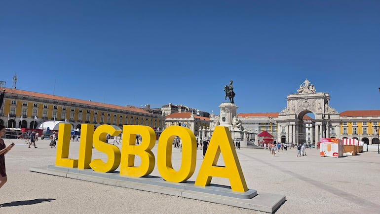 Mujer de prenda oscura tomando una fotografía junto al letrero 'LISBOA', mientras otros visitantes pasean en un día soleado.