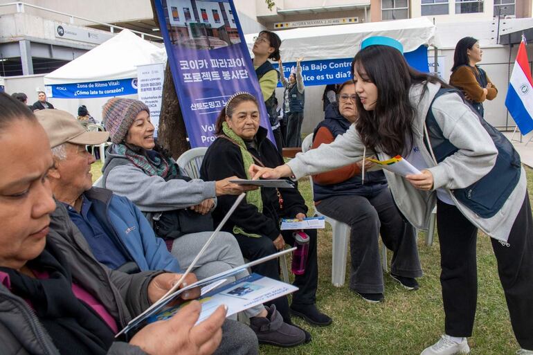 Mujer joven en chaqueta gris entrega folletos a cuatro mujeres mayores interesadas, en un ambiente tranquilo con carteles y una bandera de Paraguay.