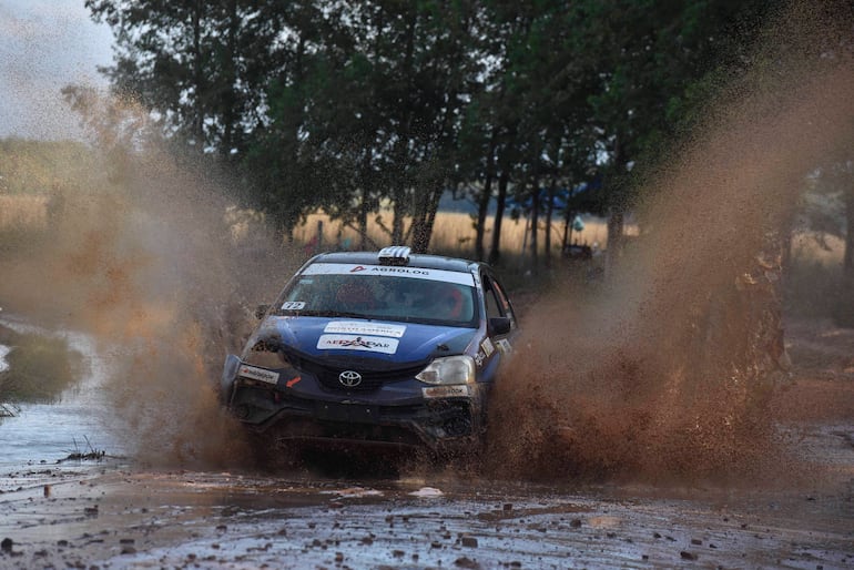 Automóvil Toyota de rally avanzando por un terreno fangoso, generando barro y agua en un paisaje natural.