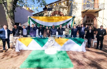 Grupo de diez personas sonrientes en evento político al aire libre, levantando las manos frente a mesa decorada con colores amarillo y verde.