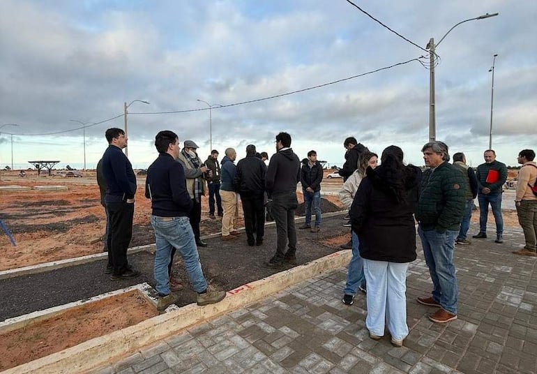 Grupo de personas vestidas formalmente discutiendo en área de construcción, con cielo nublado y farolas visibles.