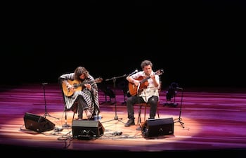Los músicos Martín Ortega y Orlando Martínez, con guitarras, lucen poncho de rayas y camisa floral en un escenario iluminado.