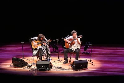 Los músicos Martín Ortega y Orlando Martínez, con guitarras, lucen poncho de rayas y camisa floral en un escenario iluminado.