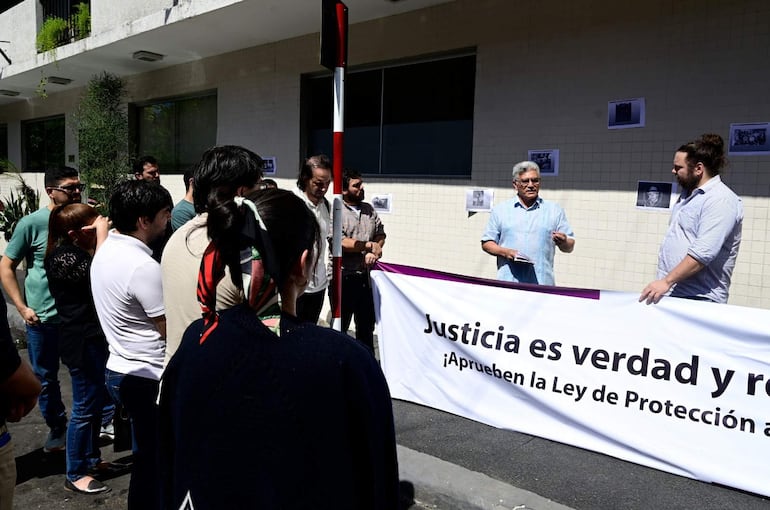 Grupo de personas frente a edificio, algunos con expresiones de tristeza, sosteniendo un cartel que dice 'Justicia es verdad'.