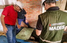 Tres hombres en un taller: uno en camiseta roja observa una tapa en el suelo, mientras otros dos realizan acciones relacionadas.