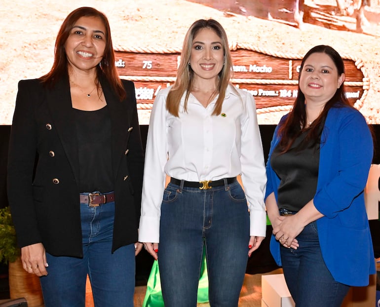 Tres mujeres sonrientes: una con abrigo negro, otra en blusa blanca y la última con chaqueta azul, conversan en un entorno decorado.