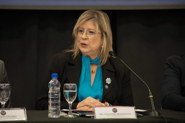Alejandra Monteoliva sentada con blusa azul y saco negro, respondiendo preguntas en panel de conferencia con documentos y vaso de agua.