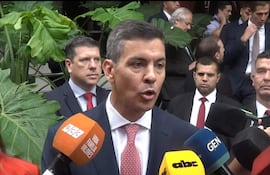 Santiago Peña con traje oscuro y corbata roja, rodeado de hombres en traje oscuro, durante una conferencia de prensa.