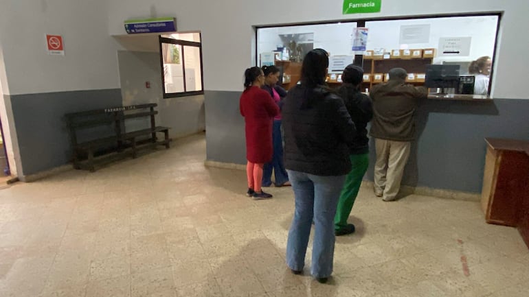 Hombre en abrigo oscuro habla con trabajadora de farmacia; mujeres en colores variados esperan en un ambiente tranquilo y ordenado.