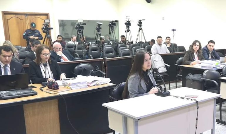 Liza Cabañas, con blusa oscura y cabello suelto, habla frente a micrófono, mientras un hombre toma notas a su lado.